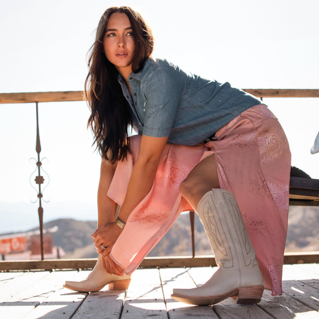 Woman standing on a wooden deck wearing pink skirt, a denim shirt, and white cowboy boots with a scenic background.