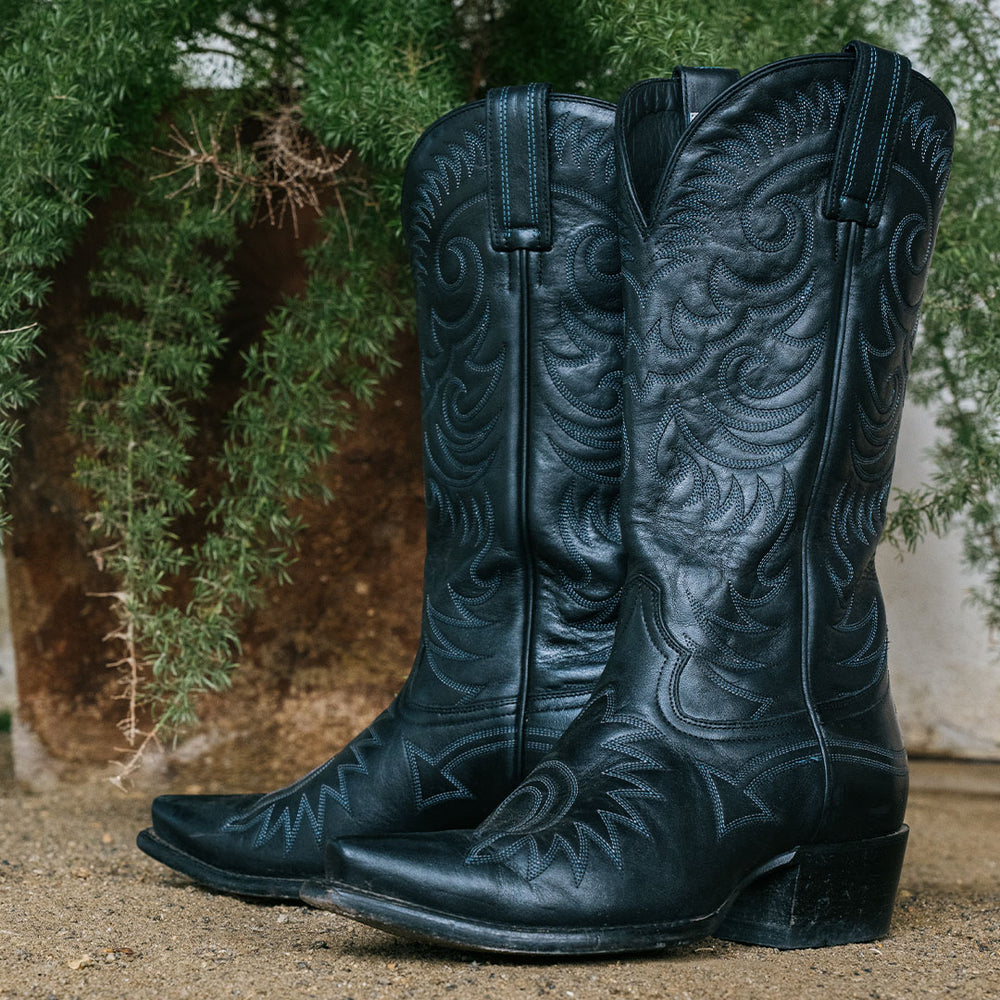 Pair of black cowboy boots with snip toe and all over stitching details sitting in front of greenery