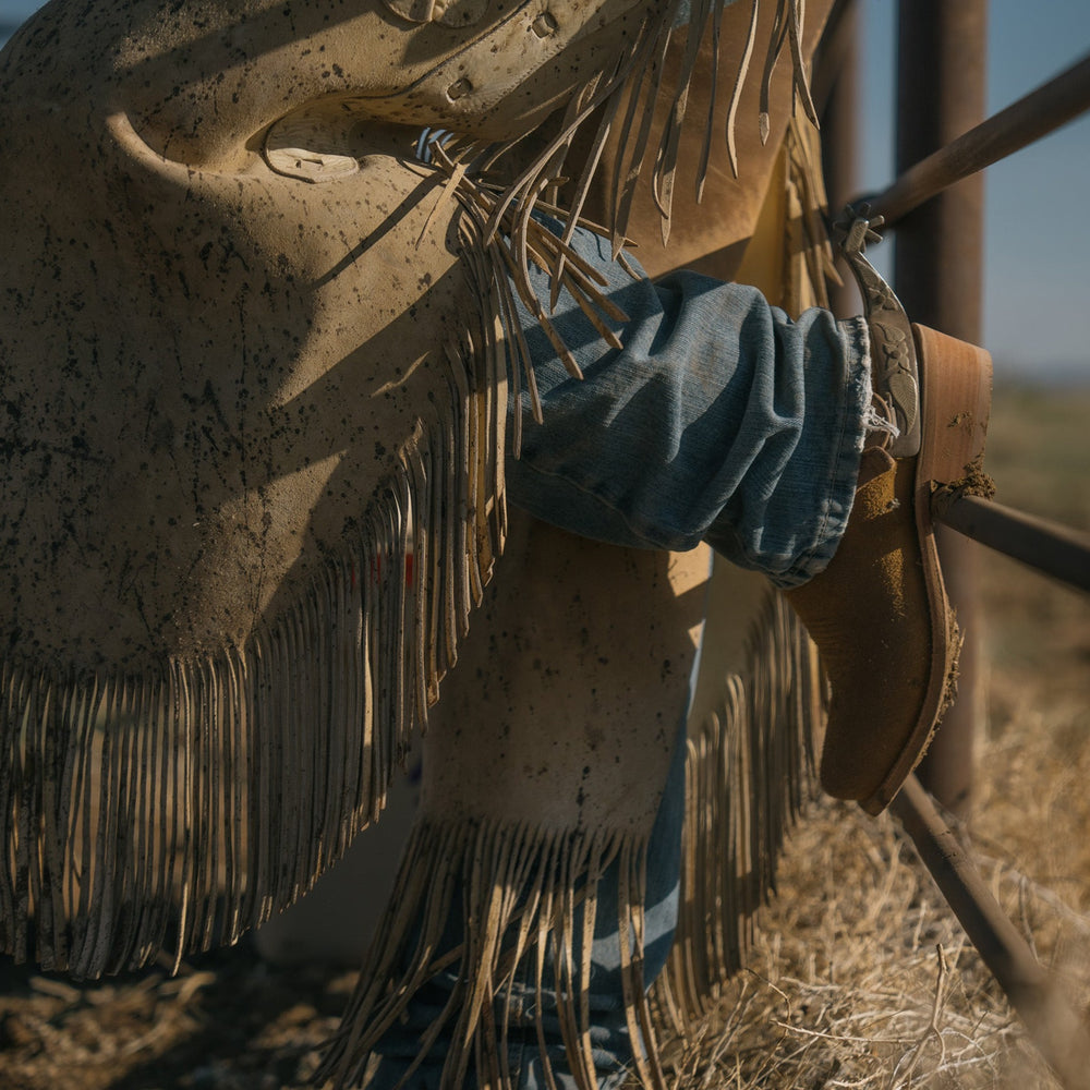 Lifestyle shot of cowboy boot resting against a metal gate, model is wearing leather fringe chaps, standing on hay