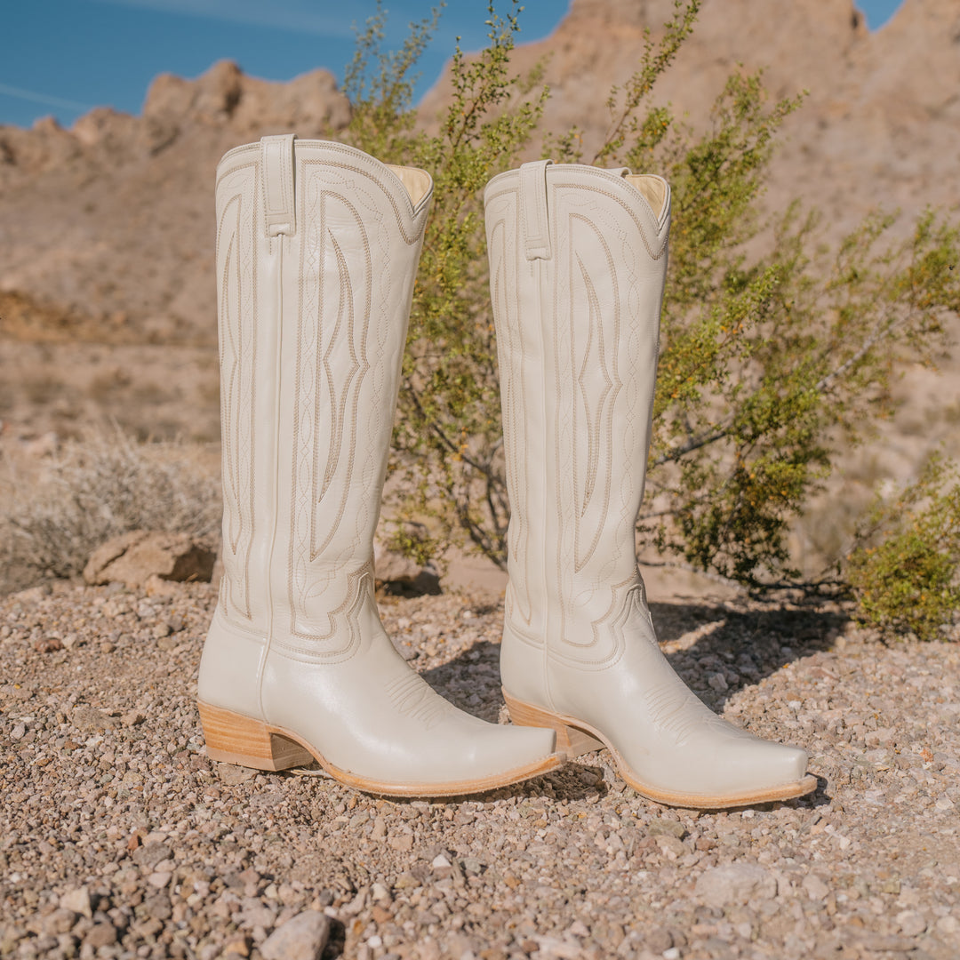 Pair of white cowboy boots on a rocky desert ground with sparse vegetation.