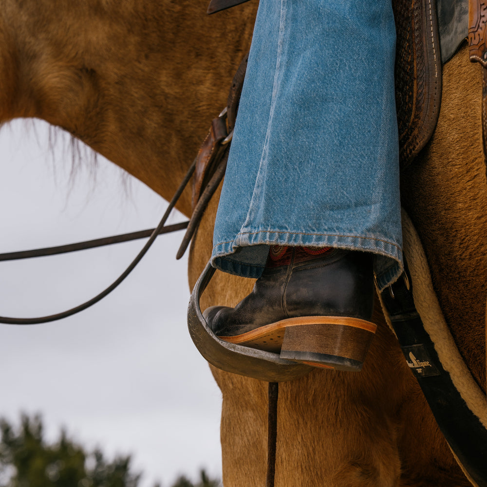 Lifestyle shot of back of cowboy boot in stirrup with rider on horse wearing blue jeans
