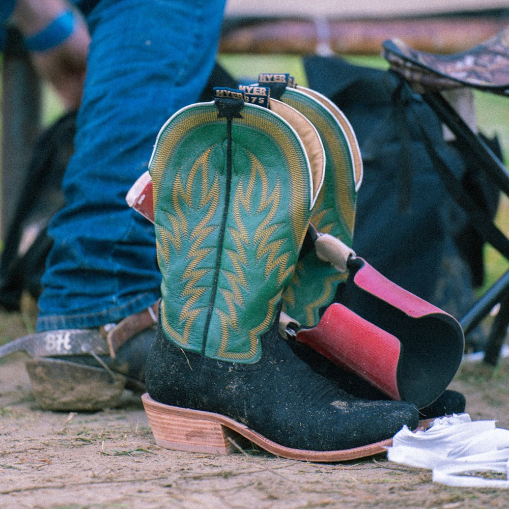 Black and green cowboy boots sitting on dirt with man standing behind them
