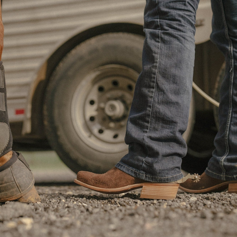 Man wearing blue jeans and brown cowboy boots with spurs standing on gravel in front of trailer