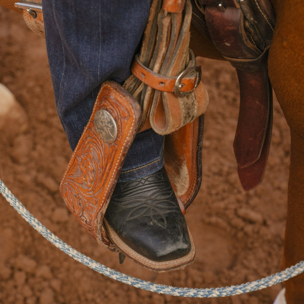 Lifestyle image of black cowboy boot in a stirrup on a dirt background