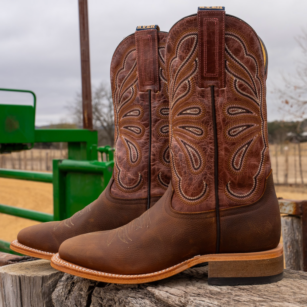 Brown cowboy boots with intricate designs on a wooden surface, with a green metal gate and blurred outdoor background.