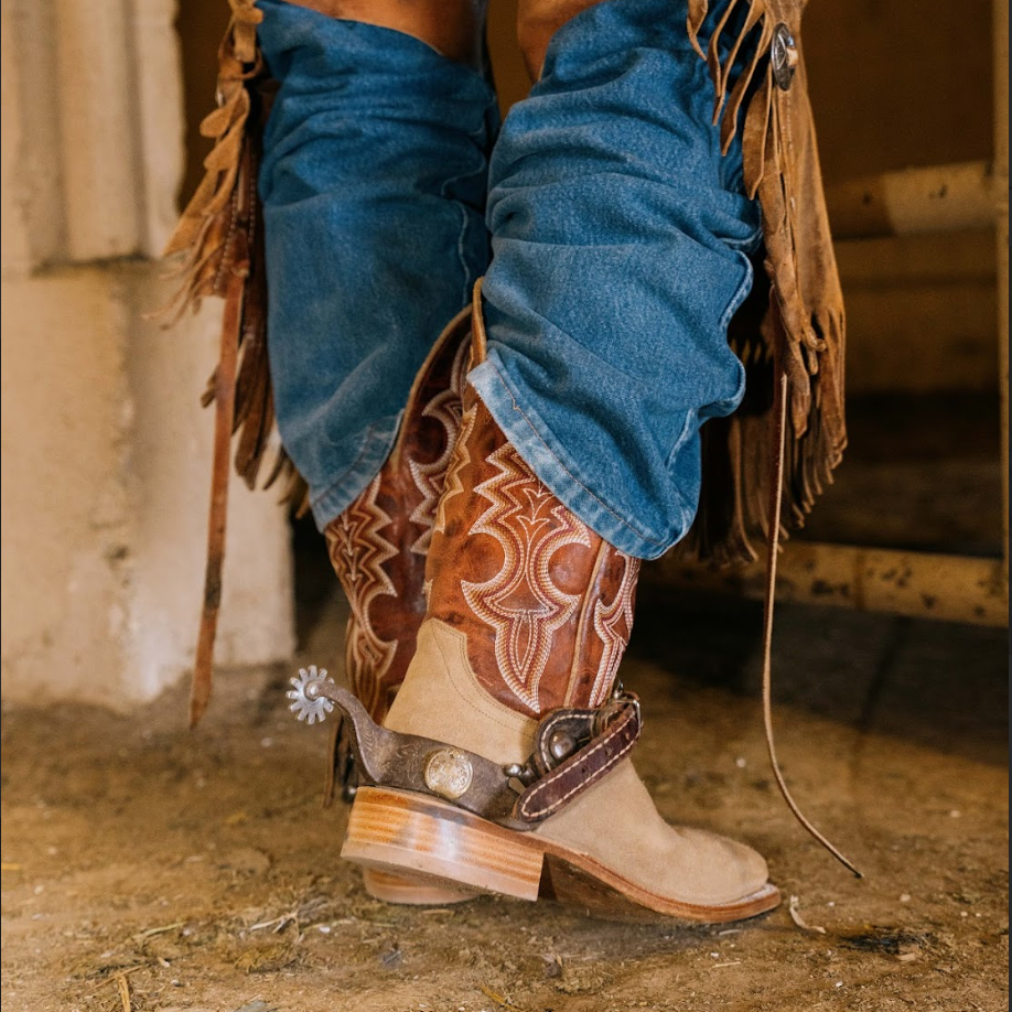 Close-up of cowboy boots with intricate stitching and spurs, worn with blue jeans in a rustic setting.